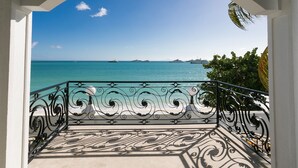 A balcony with ornate black metal railings, a clear view of the turquoise sea, and distant ships.