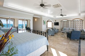 A spacious bedroom with a large bed, a balcony with a view, a seating area with sofas, and a ceiling fan.