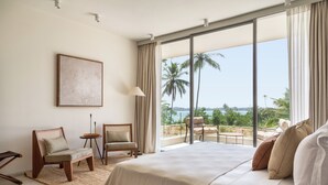 A bedroom with a large bed, two chairs, a small table, and a view of palm trees and the sea through large windows. - Tri Ahangama (Ahangama)