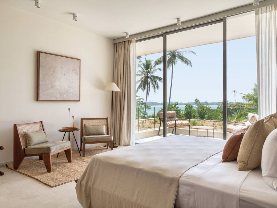 Un dormitorio con una cama grande, dos sillas, una mesita y vistas a palmeras y al mar a través de ventanas amplias.