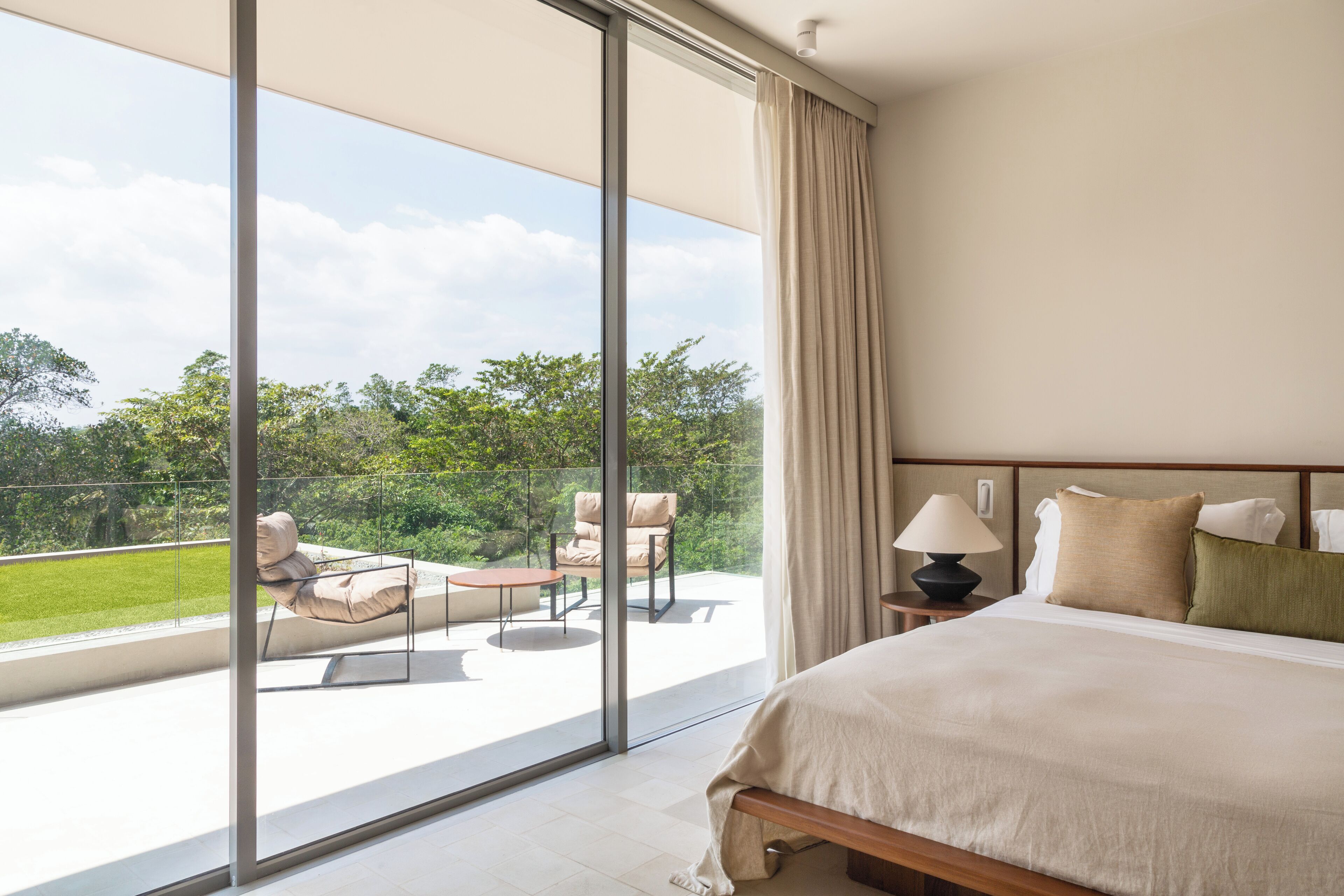 A bedroom with a large window, a bed, a nightstand, and a view of a balcony with outdoor furniture.