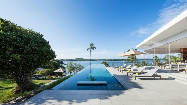 A modern infinity pool with a clear view of the lake, surrounded by lounge chairs and umbrellas. - Tri Ahangama (Ahangama)