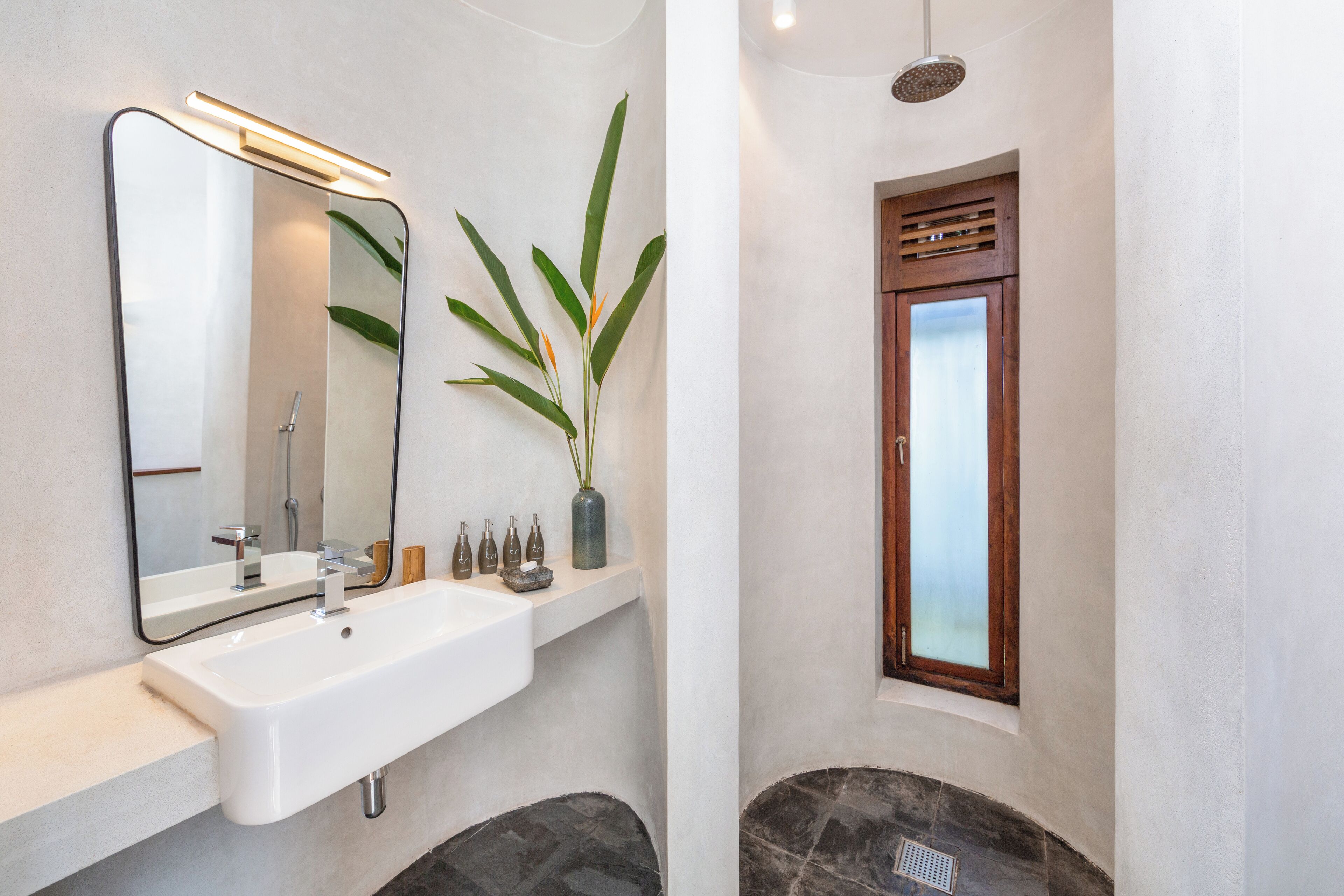 Un baño con un lavamanos blanco, un espejo, una planta en maceta y una ventana con contraventanas de madera.