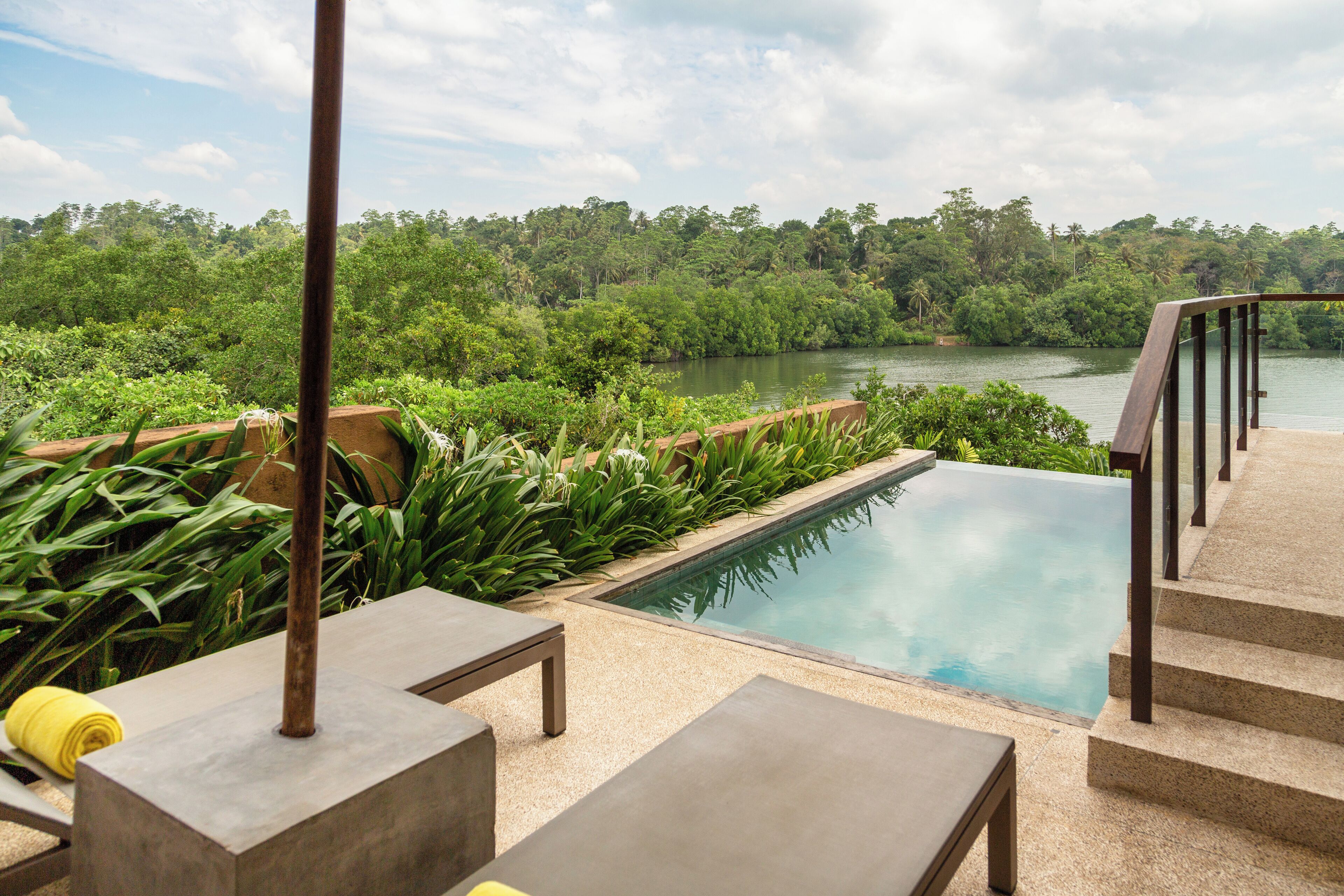 A pool with a view of a river and forest, surrounded by outdoor furniture and steps.