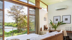 A bedroom with a large window overlooking a lake, a bed, a bedside table with a lamp, and a small potted plant. - Tri Ahangama (Ahangama)