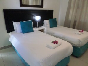 A hotel room with two beds, each with white bedding and teal pillows. There is a nightstand with a lamp between the beds. - Saldanha Bay Inn (Saldanha)