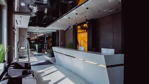 A modern hotel reception area with a glass partition, a reception desk, and a staircase leading to an upper level.