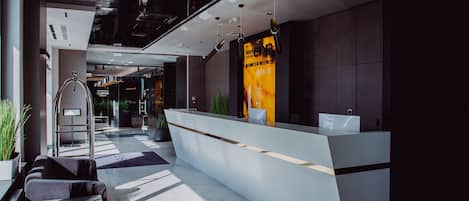 A modern hotel reception area with a glass partition, a reception desk, and a staircase leading to an upper level.