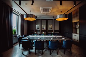 Een vergaderzaal met een lange tafel, blauwe stoelen, hanglampen en kunstwerken in lijsten aan de wanden.