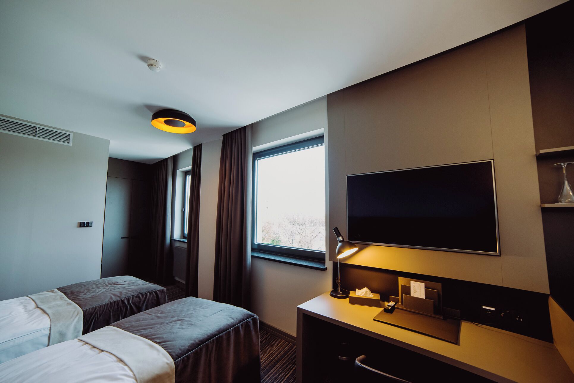 A hotel room with a bed, a desk with a flat-screen TV, a lamp, and a window.