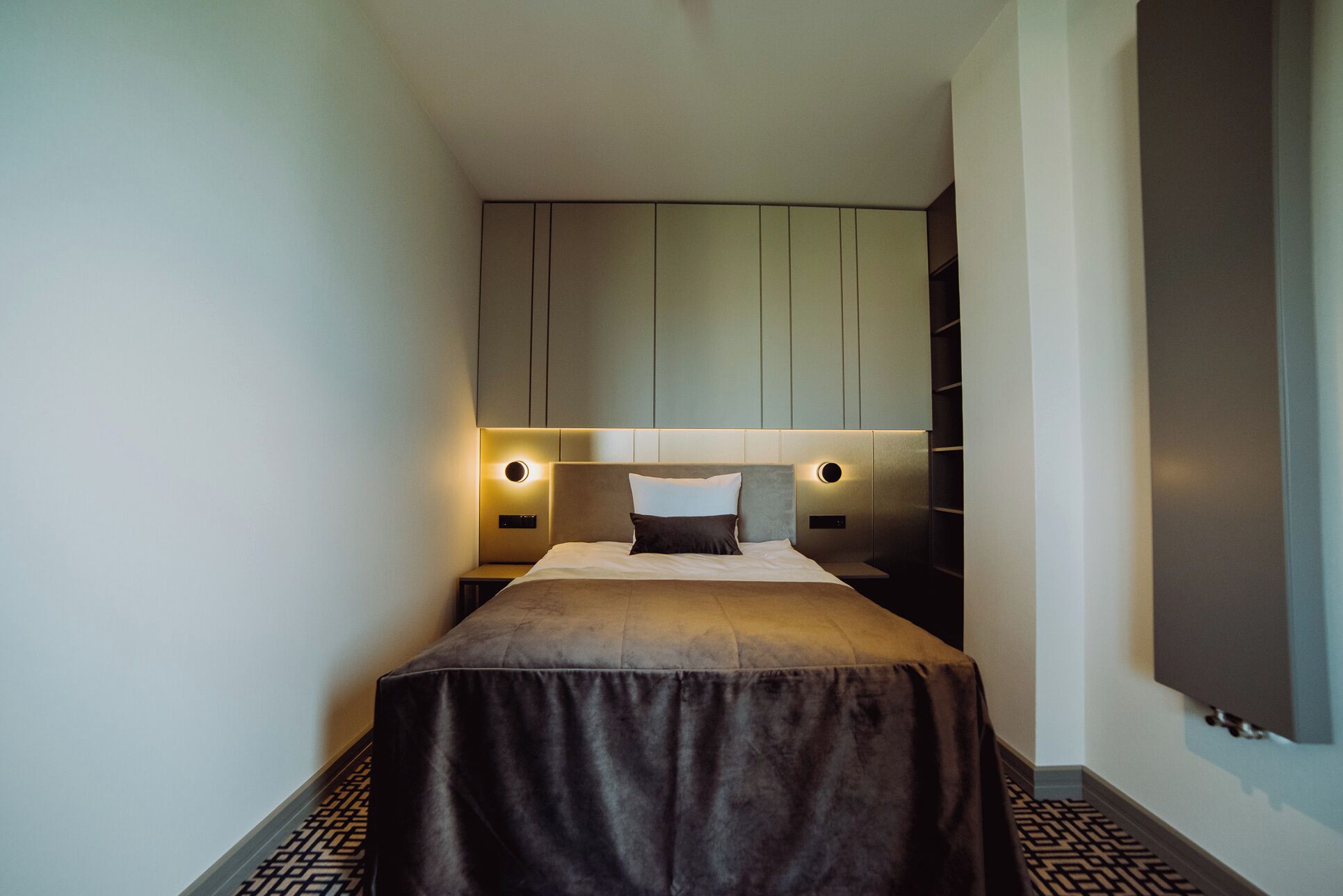 A hotel room with a bed, bedside tables, a headboard with integrated lighting, and a wardrobe.