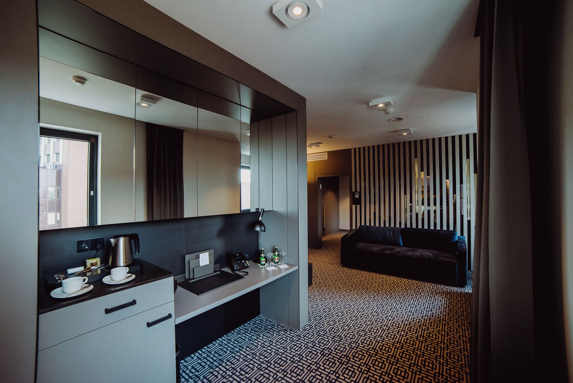 Quarto de hotel moderno com cozinha compacta, espelho grande, carpete estampado e sofá de cor escura.