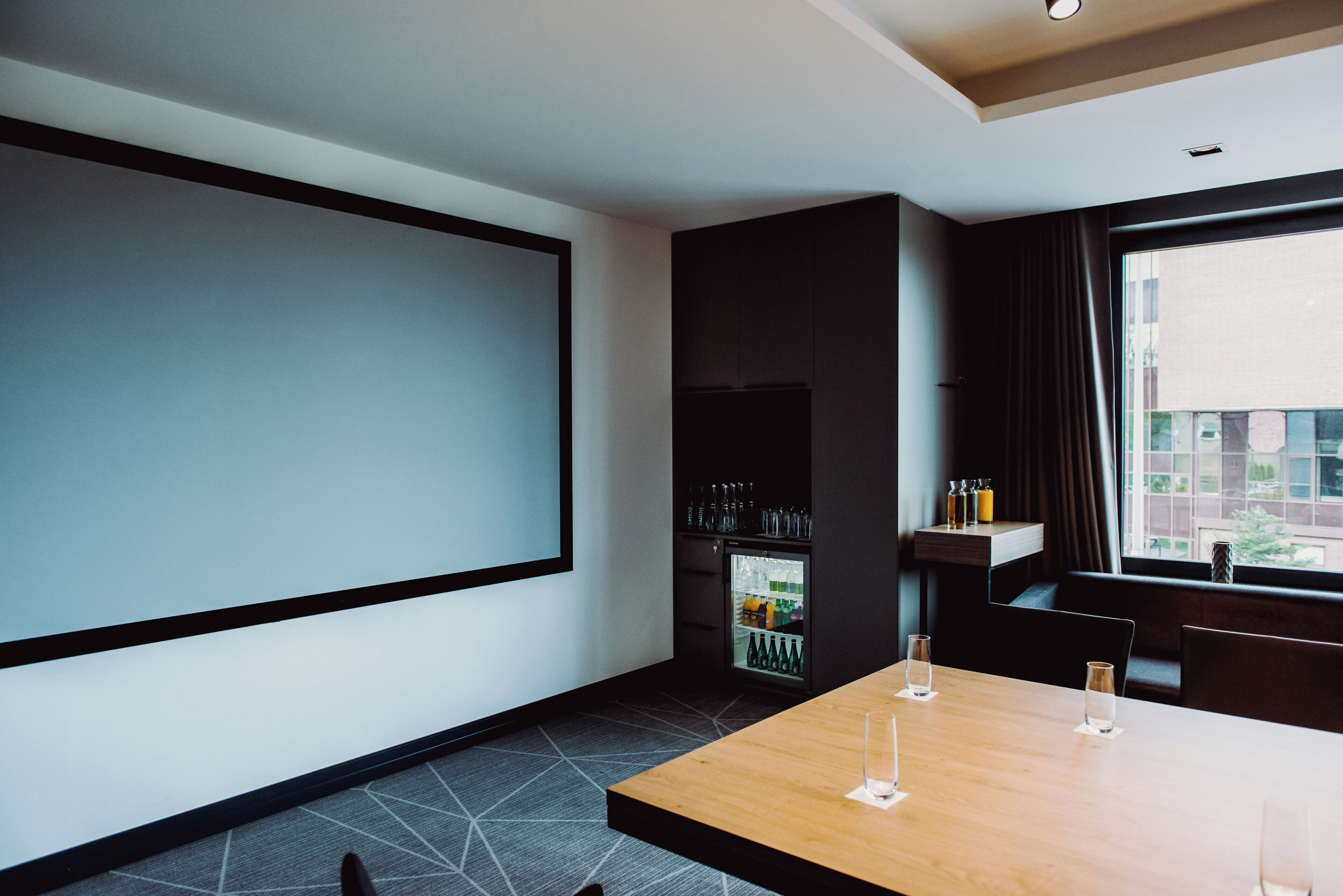 Ett modernt hotellrum med en stor tv-skärm, ett träbord med glas, en minibar och utsikt över byggnader genom fönstret.