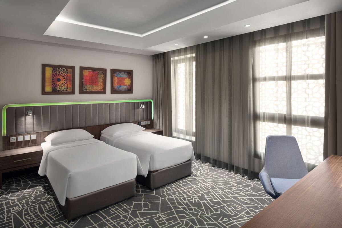 A hotel room with two beds, a desk, and a chair. There are three artworks on the wall, and the room has a large window with curtains.