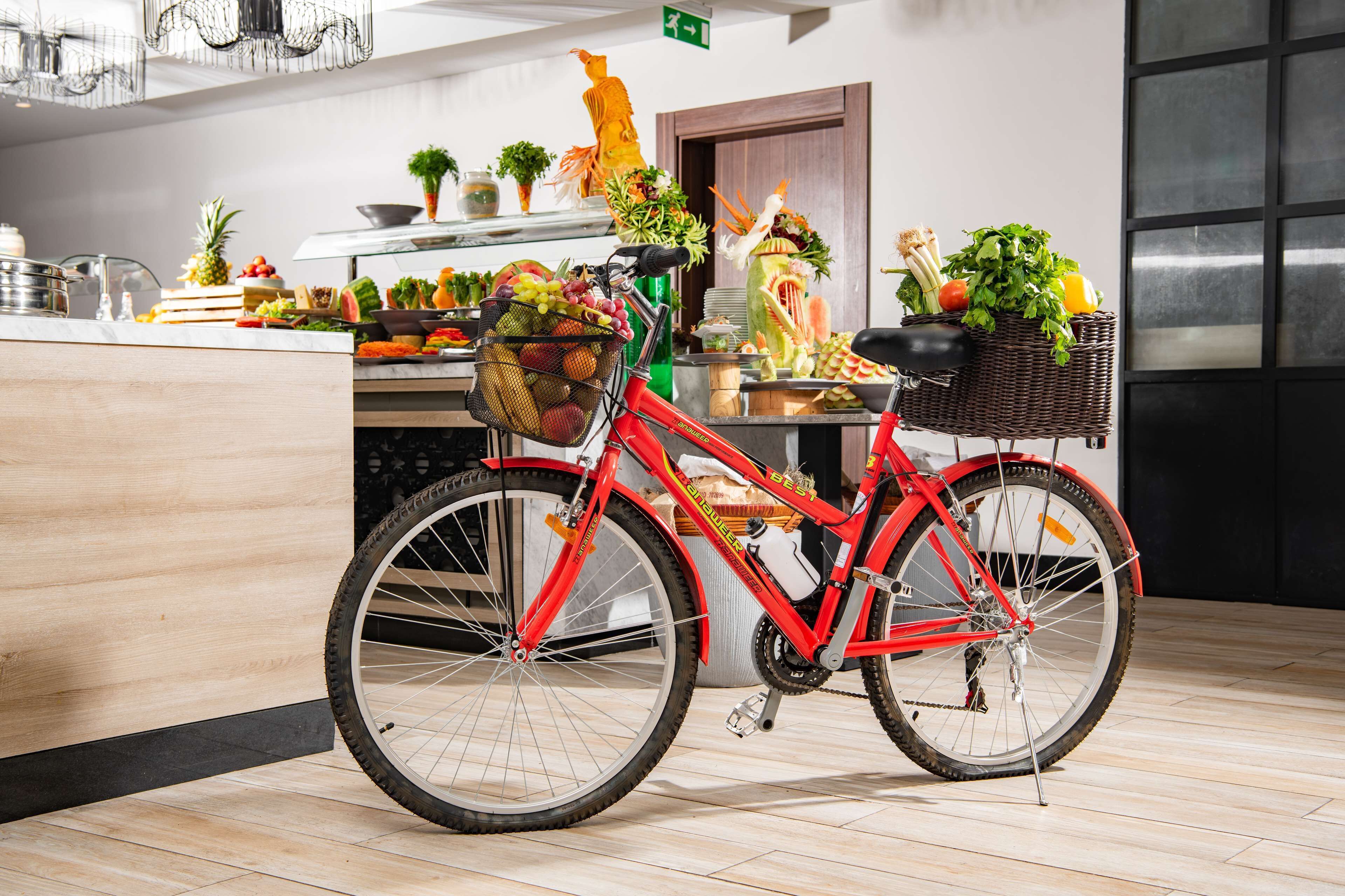 A red bicycle with a basket, parked in a modern kitchen with wooden flooring and various fruits and vegetables displayed on the counter.