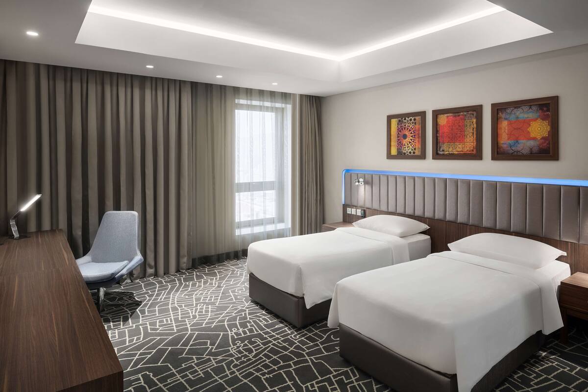 A hotel room with two beds, a desk, a chair, and a large window with curtains.