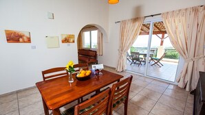 Ein Essbereich mit Holztisch und Stühlen, einer Vase mit Blumen und einer Schüssel Orangen. Ein Fenster mit Vorhängen bietet Blick ins Freie.