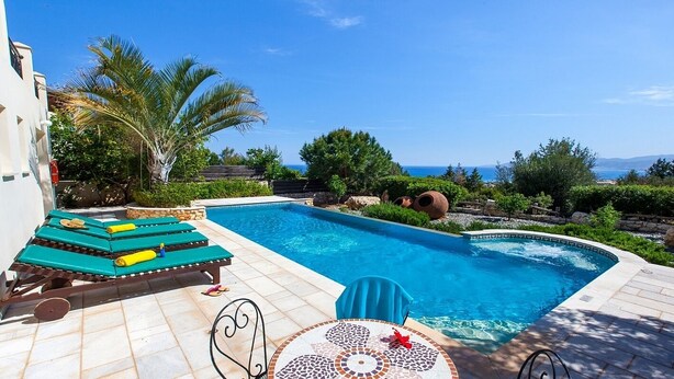 Ein Schwimmbecken mit Liegestühlen und einem Tisch, umgeben von Grünpflanzen und Blick aufs Meer.