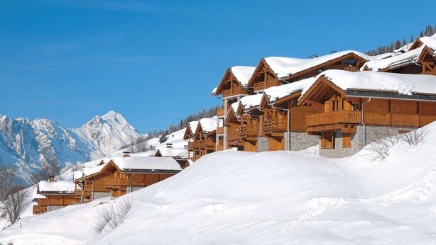 A row of wooden chalets with snow-covered roofs, set against a backdrop of snow-covered mountains and a clear blue sky. - Résidence Odalys Le Grand Panorama 1 (Valmeinier)