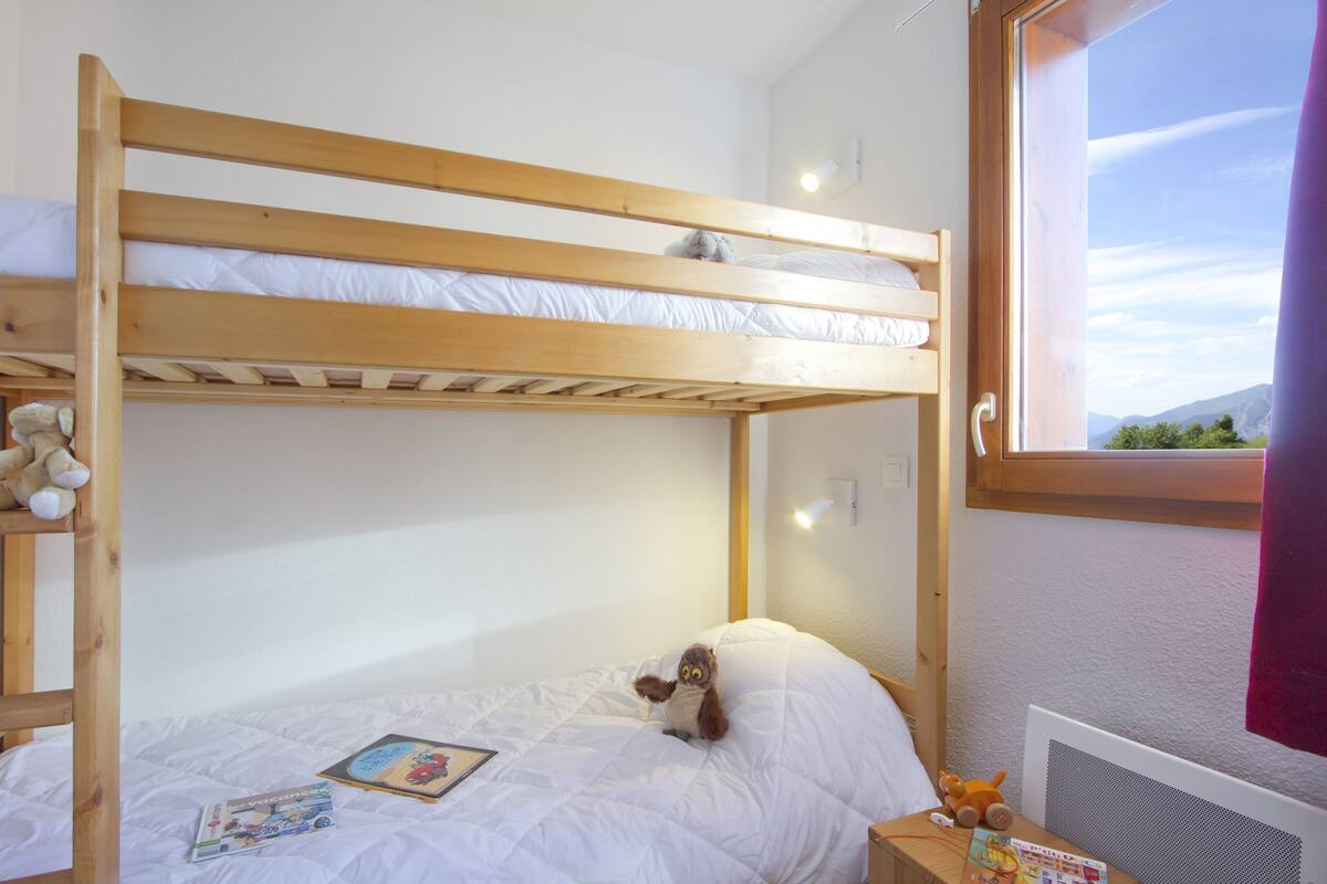Een slaapkamer met een stapelbed, een raam, een teddybeer en een pluchen dier op het onderbed.