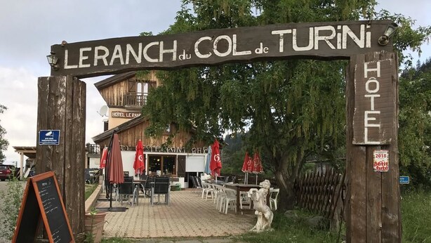 A wooden sign with "LERANCH du COL de TURINI HOTEL" written on it, leading to a building with outdoor seating and a statue. - Hotel Restaurant Le Ranch de Turini (La Bollene-Vesubie)