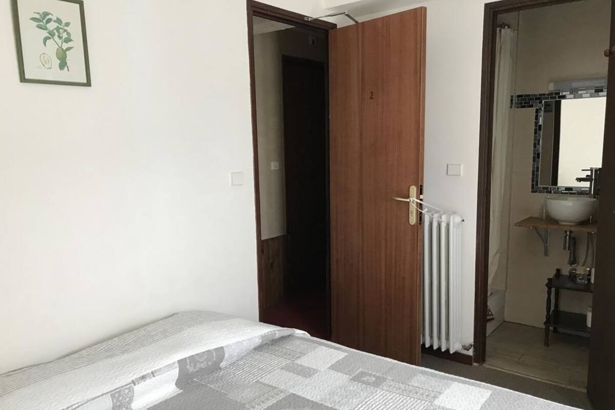 Une chambre à coucher avec un lit, une porte en bois, un radiateur et une salle de bain visible à travers une porte ouverte.