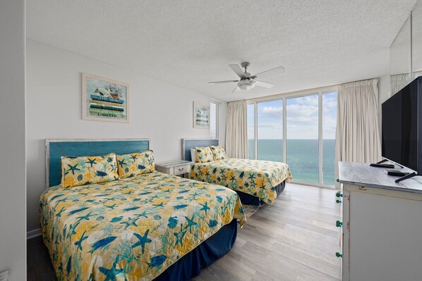 Habitación de hotel con dos camas, ventilador de techo, ventana grande con vista al mar y escritorio con televisión.