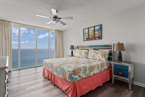 A bedroom with a large bed, a ceiling fan, a nightstand with a lamp, and a view of the ocean.