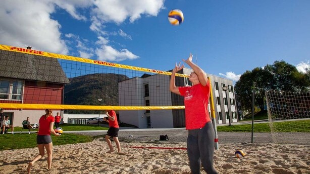 A volleyball game on a sandy court with a net, buildings, and a clear sky in the background. - Førde Sommarhotell (Sunnfjord)