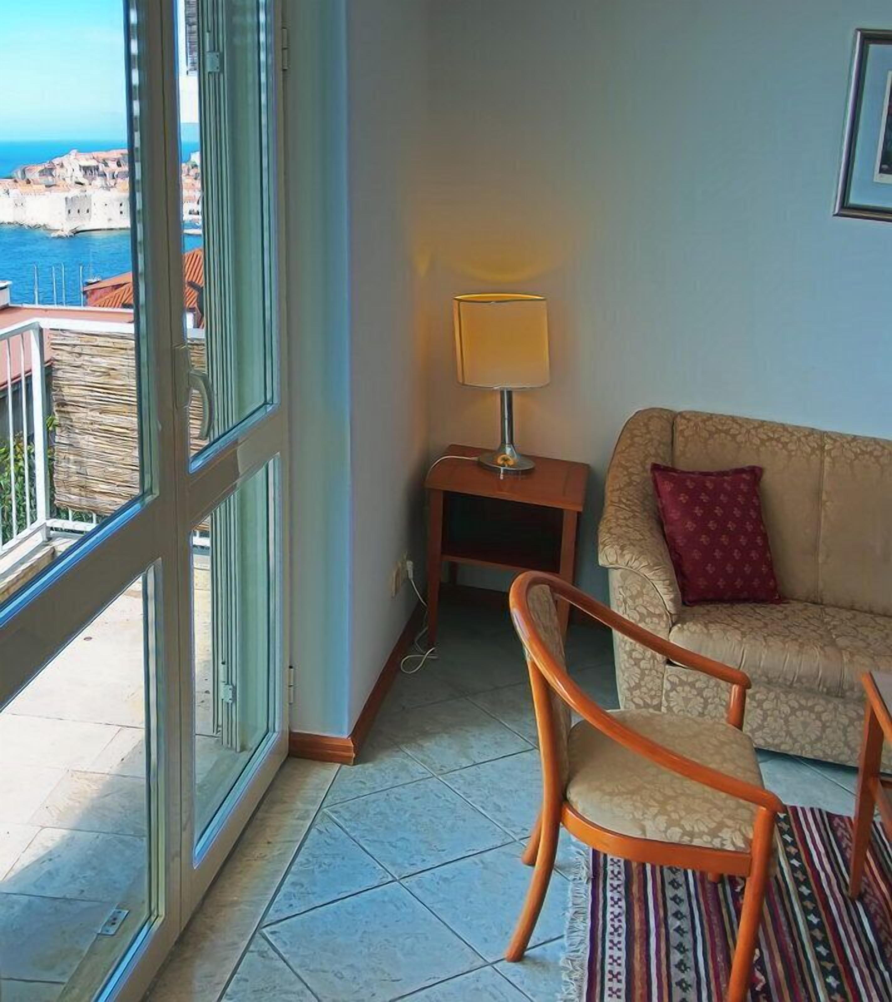 A balcony with a view of a coastal town, a small table with a lamp, a sofa with a patterned cushion, and a wooden chair.
