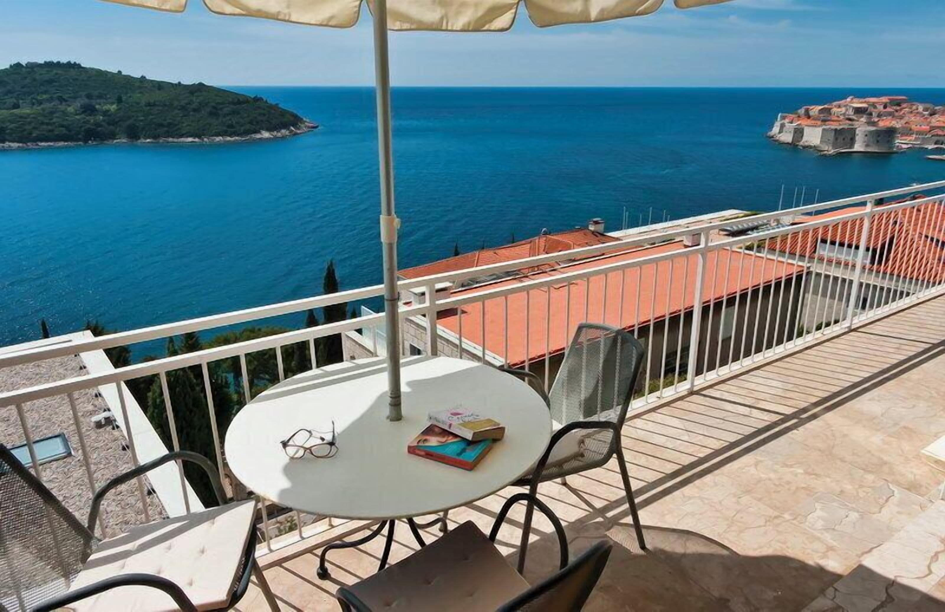 A balcony with a table, chairs, and an umbrella overlooking a coastal view with a distant island and a fortified town.