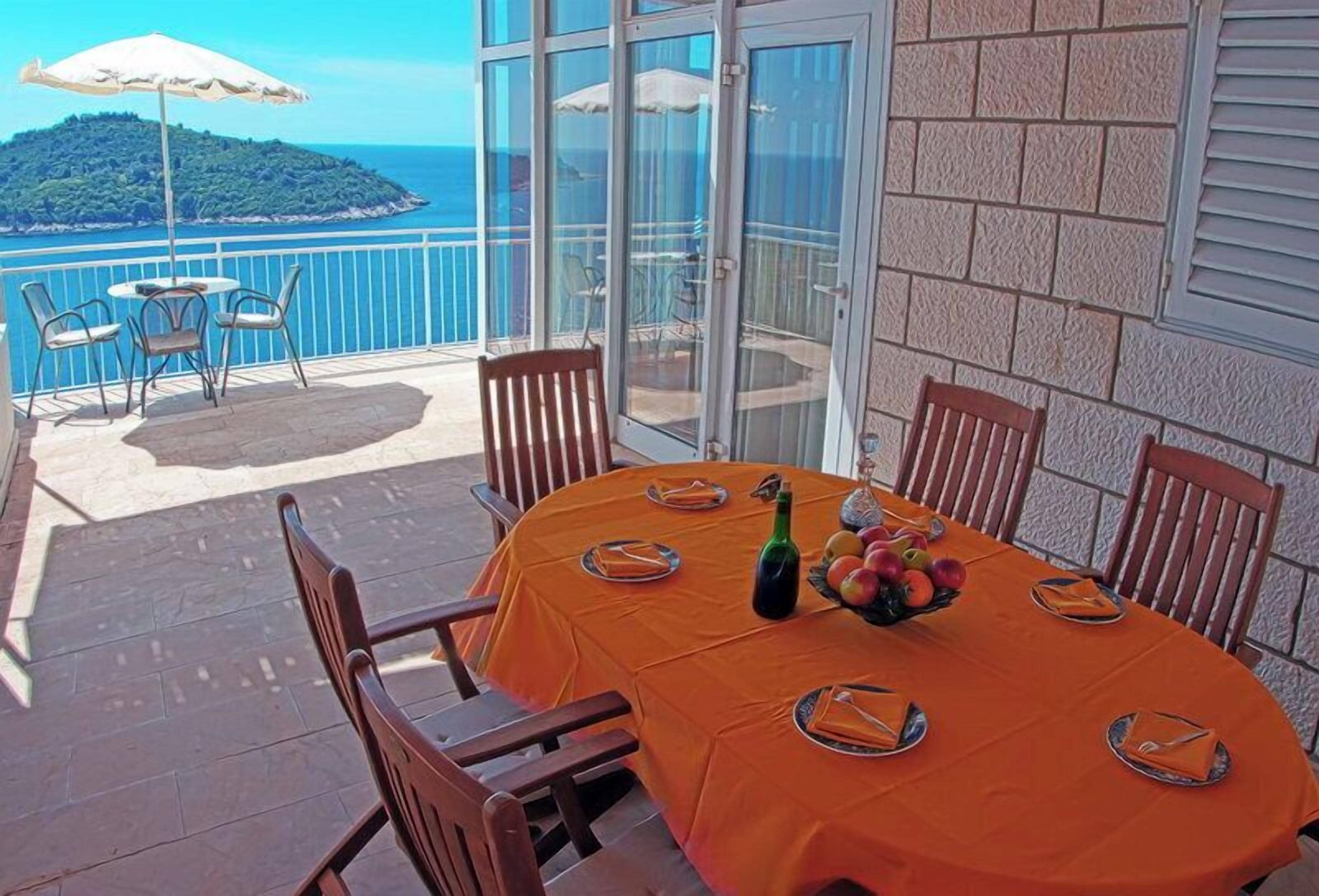 A round table with an orange tablecloth, surrounded by wooden chairs, set on a balcony with a view of the sea and a distant island.