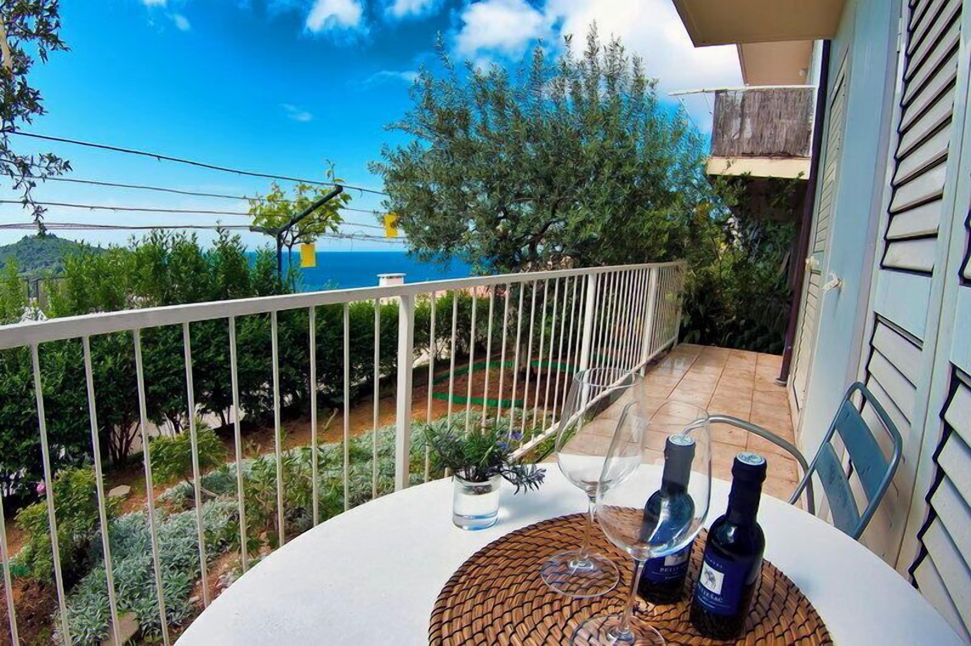 A balcony with a view of the sea, a table with a bottle of wine, and a small plant.