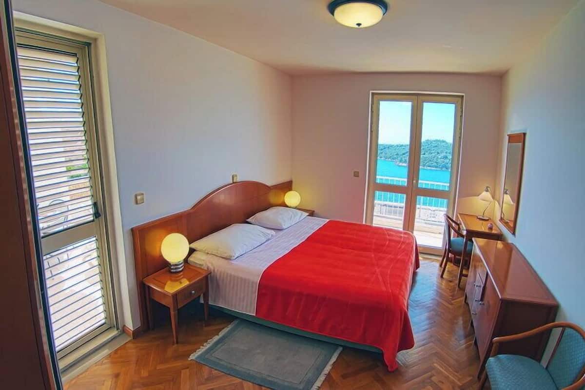 Ett sovrum med en säng, sänglampor, ett skrivbord och en stol. Det finns ett fönster med persienner och utsikt över havet.