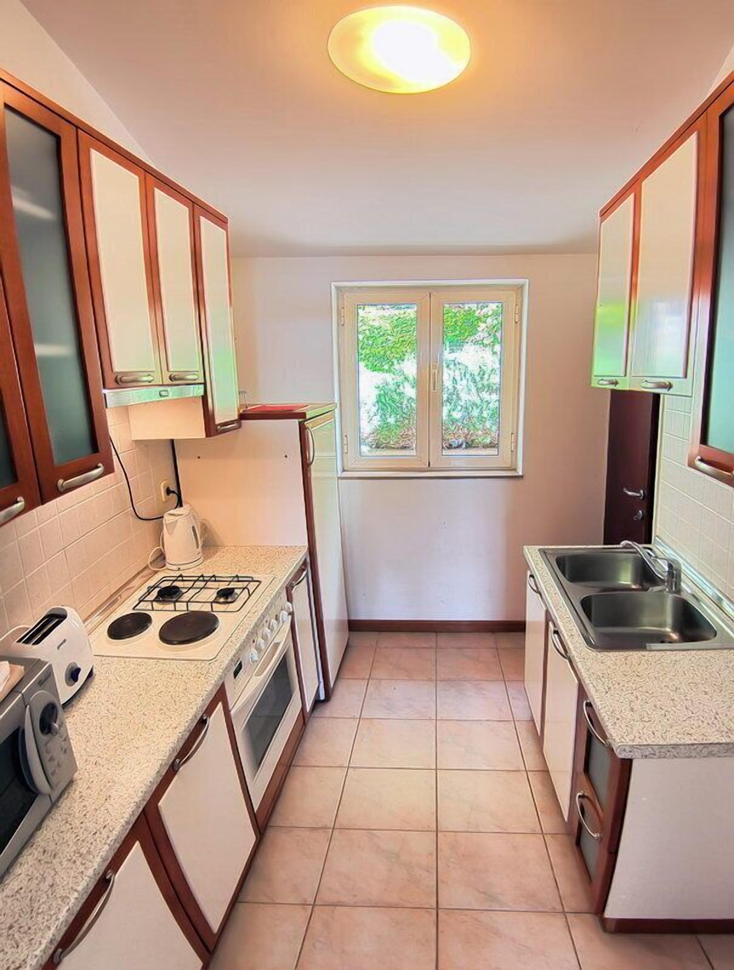 Une cuisine de taille réduite, équipée d’une cuisinière à gaz, d’un évier et d’une fenêtre donnant sur un espace verdoyant.