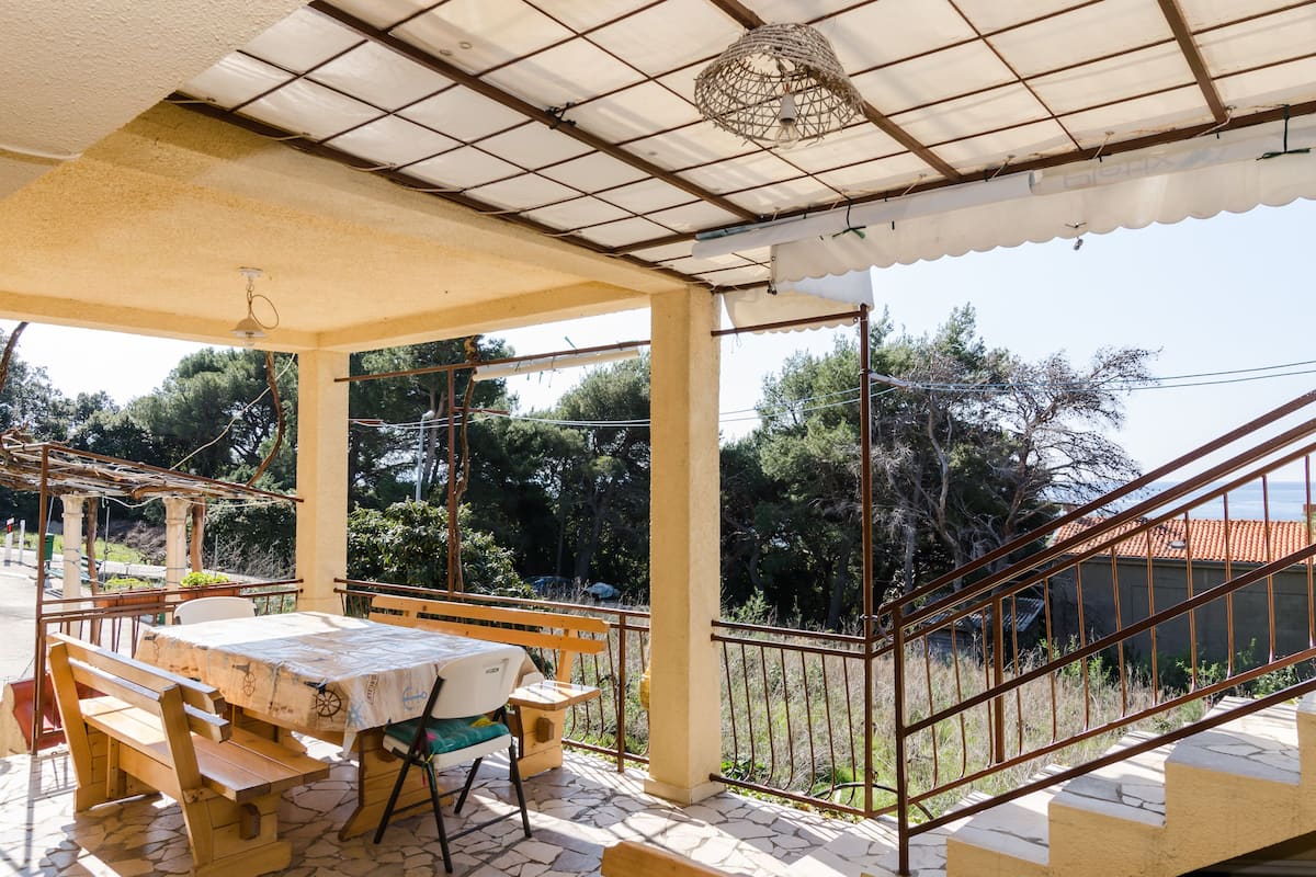 Une terrasse extérieure couverte, dotée d’une table et de bancs, qui offre une vue panoramique sur un paysage verdoyant, avec des arbres et un bâtiment en arrière-plan.