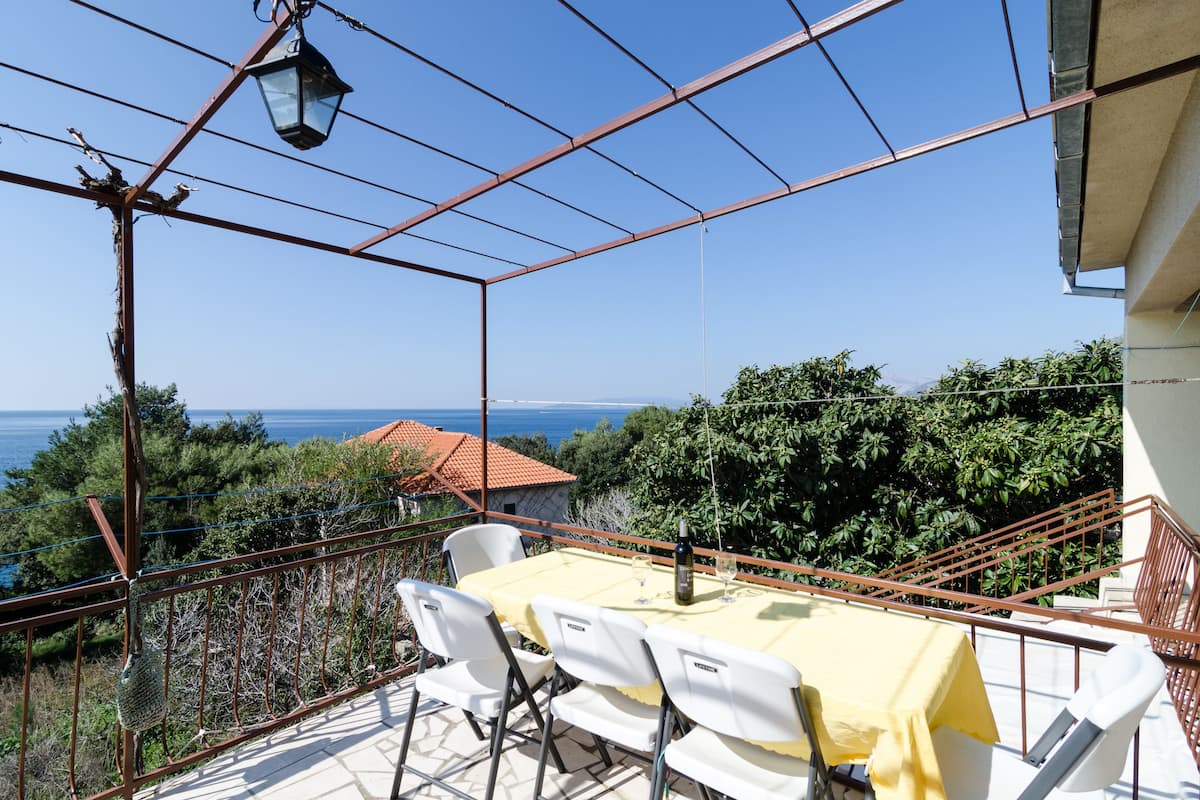 Un balcon avec une table dressée pour deux personnes, offrant une vue panoramique sur la mer et la verdure.