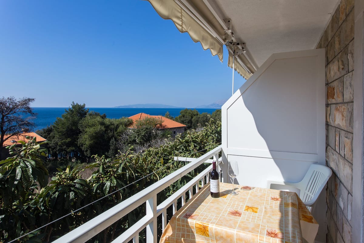 Un balcon avec une table, une bouteille de vin et un verre. Le balcon donne sur une zone côtière, avec des arbres et des bâtiments.