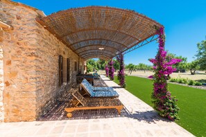 Un edificio de piedra con un espacio de estar al aire libre cubierto, que incluye tumbonas de madera y una pérgola.