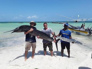 Three individuals holding large fish on a sandy beach with boats in the background.