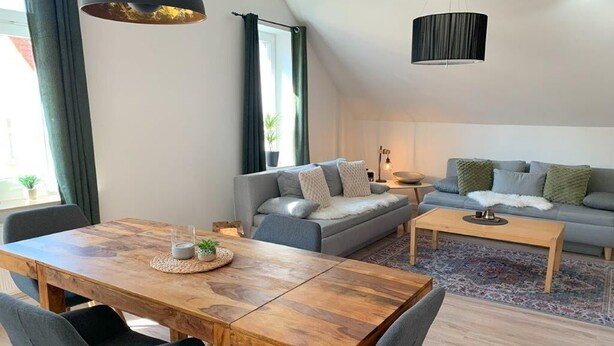 A modern living room with a wooden dining table, grey sofas, and a large window with curtains.