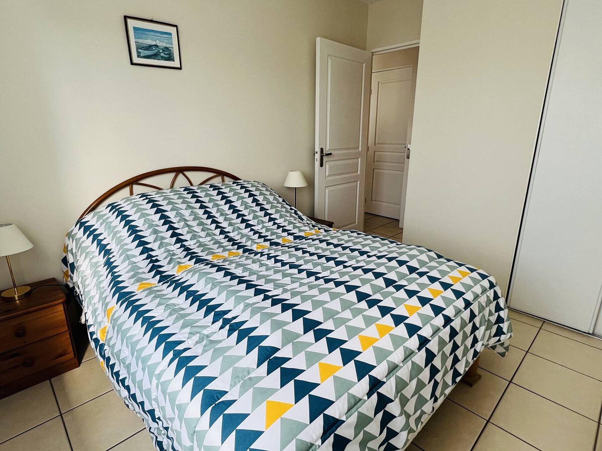 Una cama bien hecha con una colcha de estampado geométrico, una mesita de noche con lámpara y una puerta al fondo.