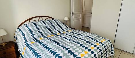 Una cama bien hecha con una colcha de estampado geométrico, una mesita de noche con lámpara y una puerta al fondo.