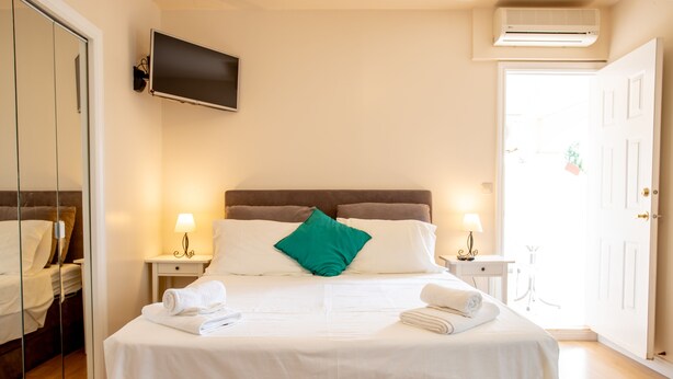 Una cama bien tendida con sábanas blancas y un almohadón verde, lámparas de noche, un televisor de pantalla plana en la pared y una puerta blanca con una planta colgando.