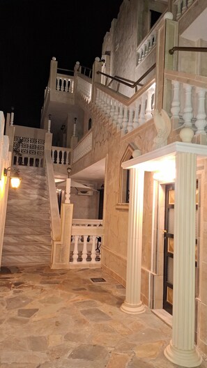 Eine imposante Treppe mit aufwendiger Balustrade führt zu einem Balkon im zweiten Stock, umgeben von Steinmauern und Säulen.