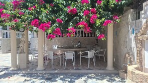 A covered outdoor seating area with a round table and chairs, surrounded by a stone wall and blooming pink flowers. - Castellino Studios - Twin Studio 4 (Faliraki)