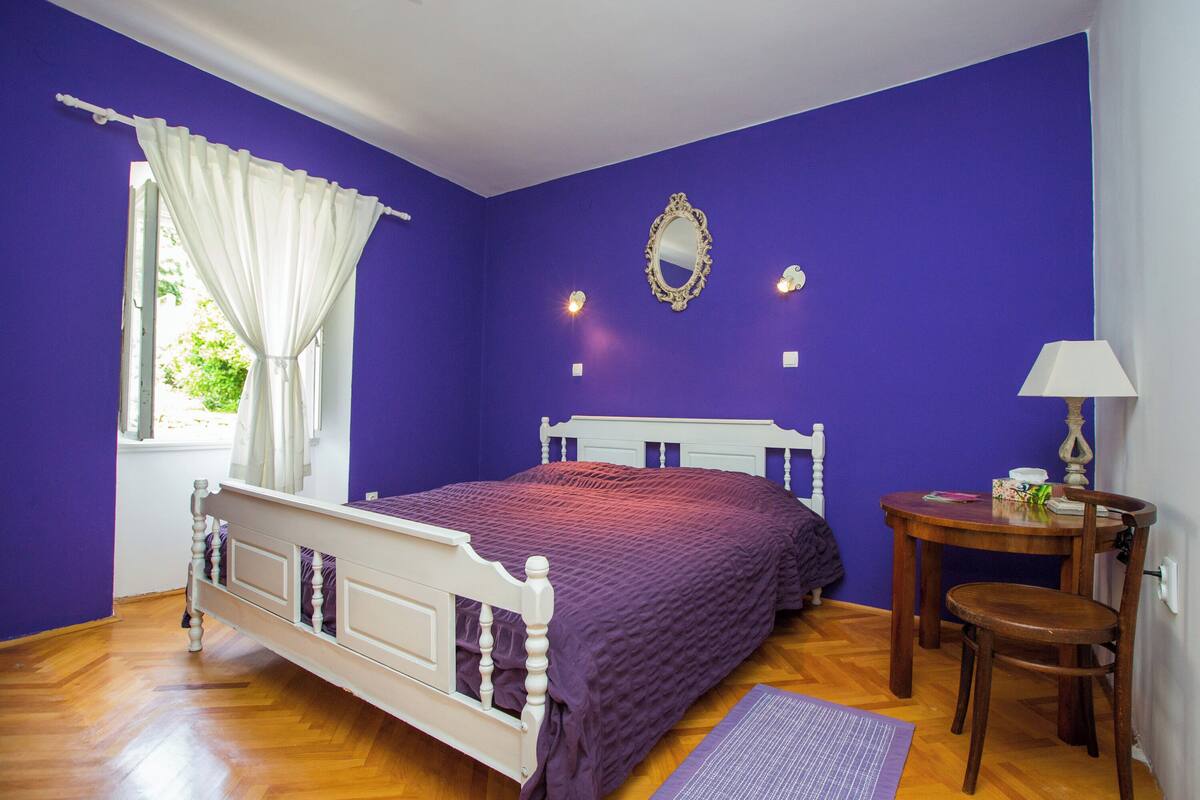 A bedroom with a bed, a chair, a table, a mirror, and a window with curtains.