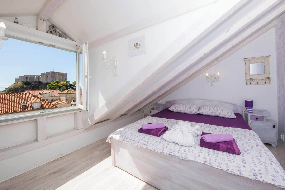 A bedroom with a bed, a nightstand, a window, and a view of a castle.