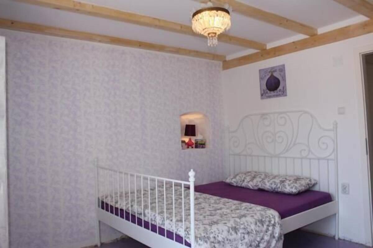 A bedroom with a white metal bed frame, a purple mattress, and a patterned bedspread. There is a small shelf with books and a lamp on the wall.