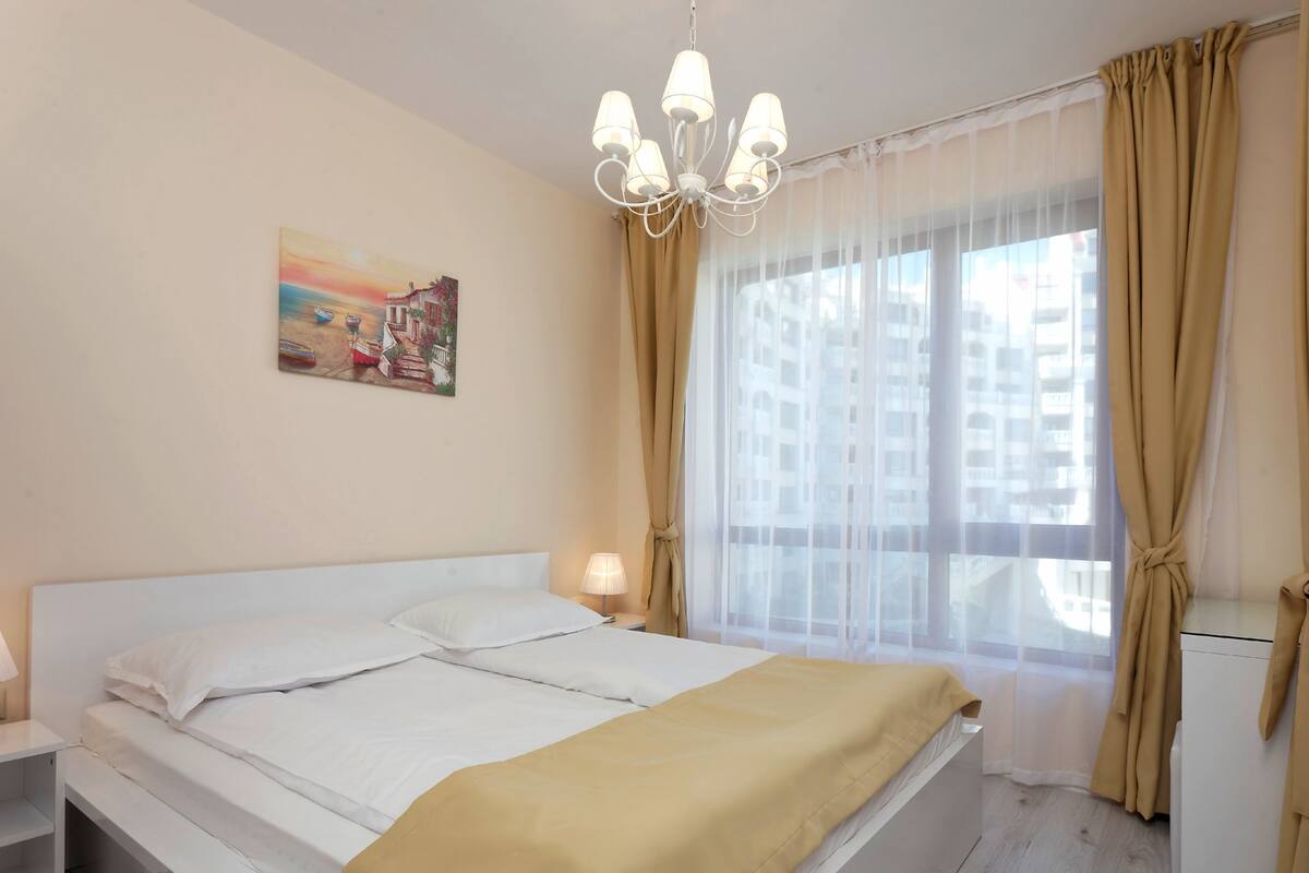 A hotel room with a bed, a bedside lamp, a painting on the wall, and a window with curtains.